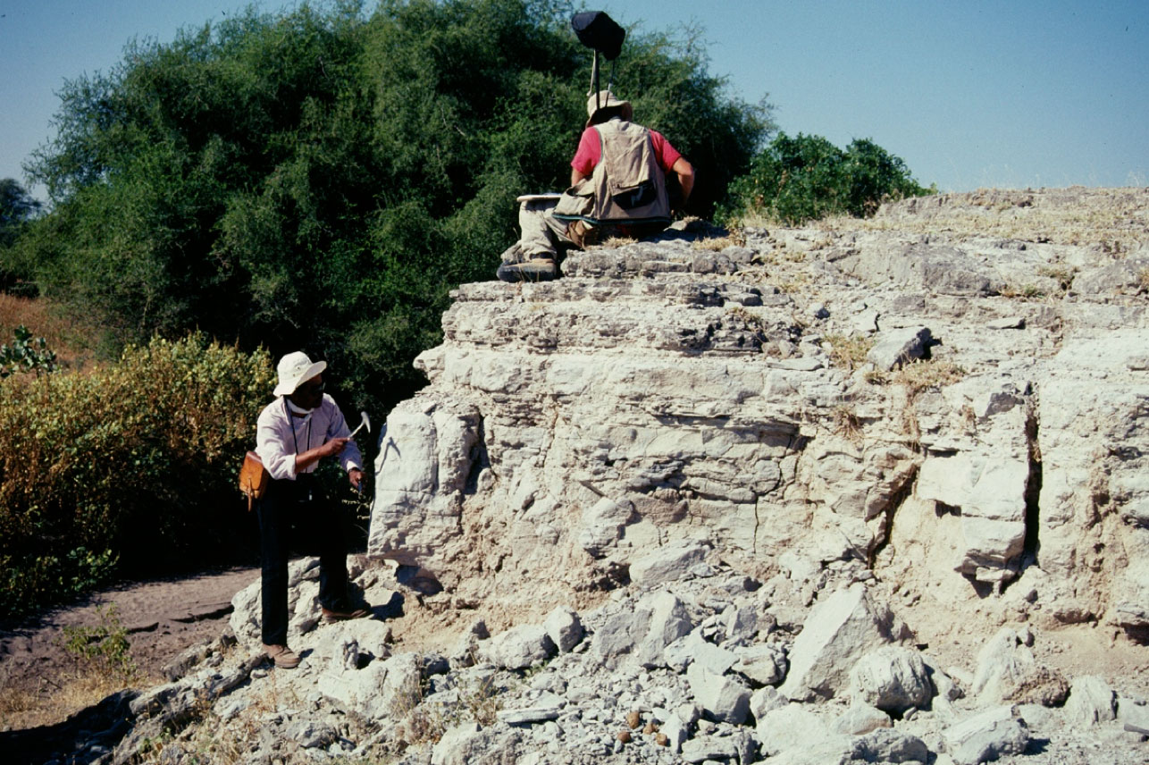 A person standing next to a rock formation with a rock hammer and a person sitting on the rock formation working.