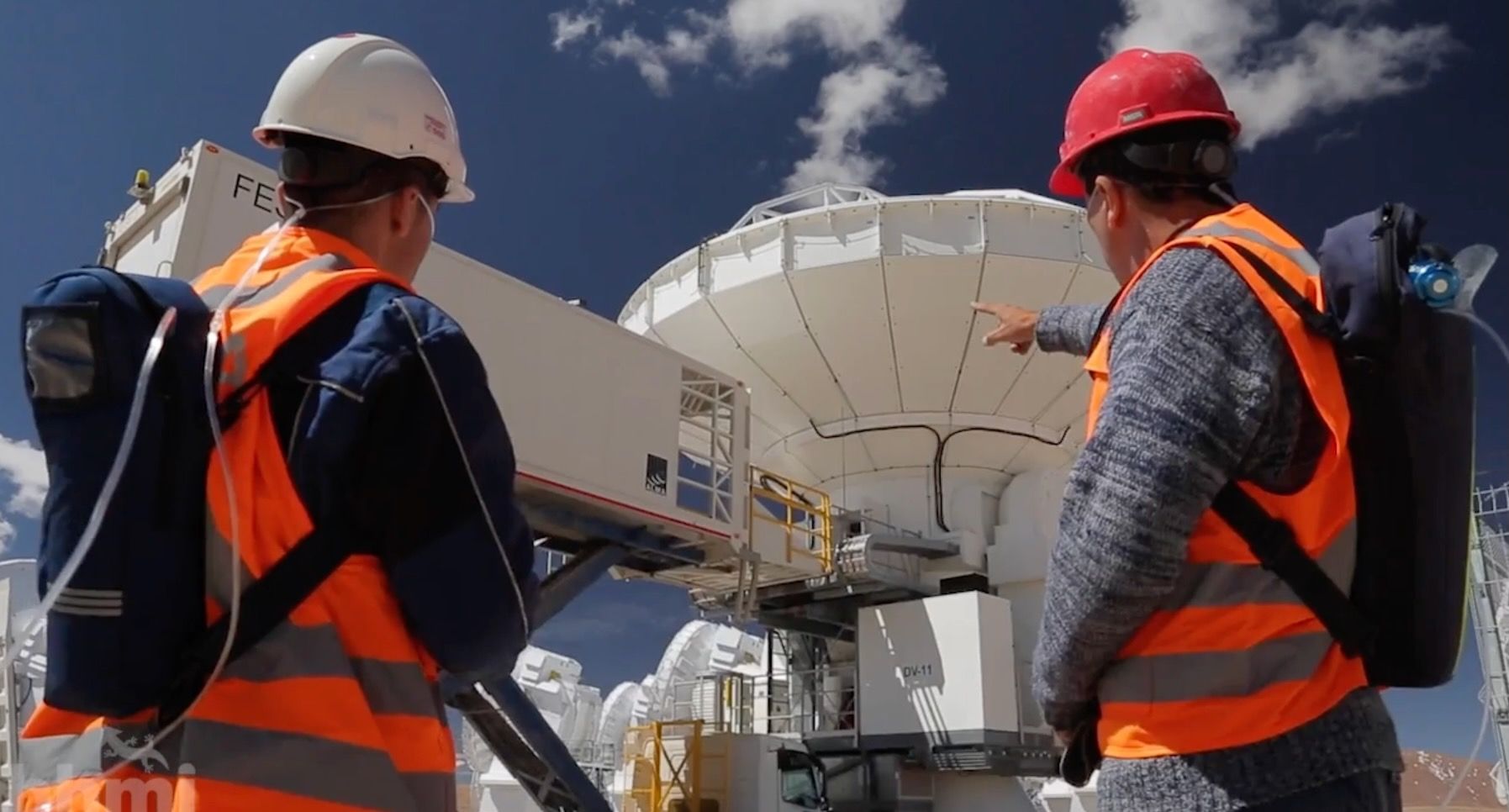 Fotografía de dos trabajadores con mochilas con oxígeno suplementario, de pie frente a un conjunto de antenas de que se encuentran a gran altitud y señalándolas.