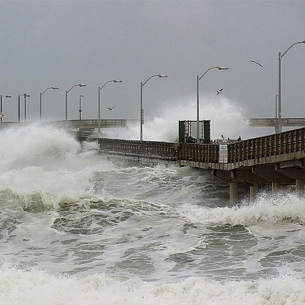 A photograph of large waves crashing against a pier on a gray stormy day.